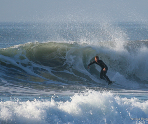A guy in a wetsuit surfing a big, cold-looking wave in the afternoon. Photo credit: Patty Bruno.